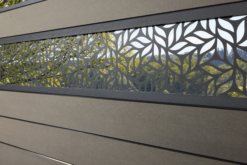 Moderner Sichtschutzzaun mit dekorativer, durchbrochener Aluminium-Ornamentleiste vor grünem Landschaftshintergrund.