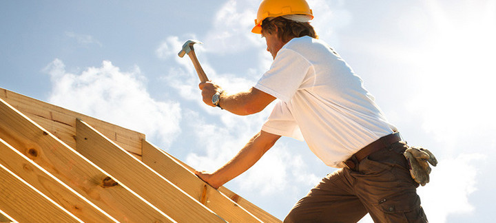 Dachdecker mit gelbem Schutzhelm beim Befestigen von Konstruktionsvollholz (KVH) auf einem Dachstuhl unter strahlendem Himmel.