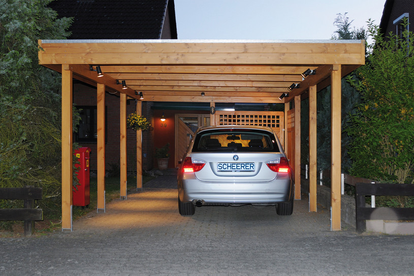 Holz-Carport bei Dämmerung mit Beleuchtung, geparktem Auto und bepflanztem Vorgarten.
