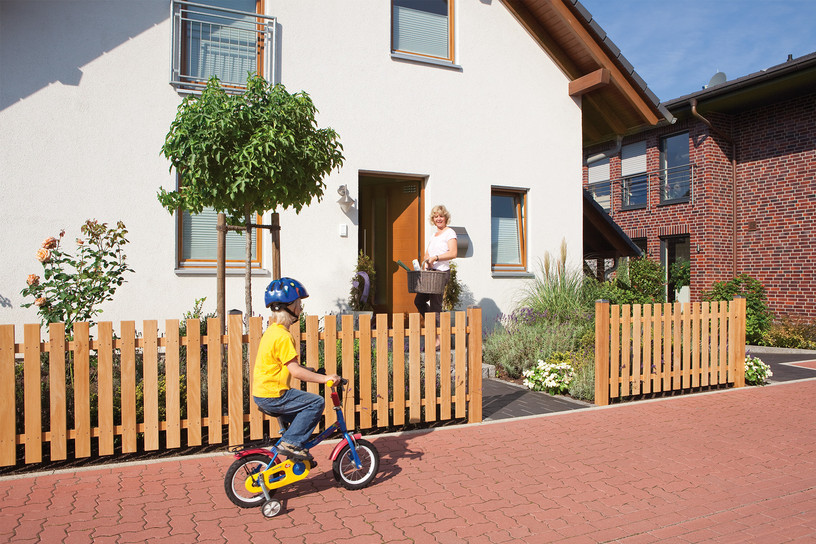 Holz-Vorgartenzaun vor Einfamilienhaus, spielendes Kind auf Fahrrad, Frau an der Haustür.