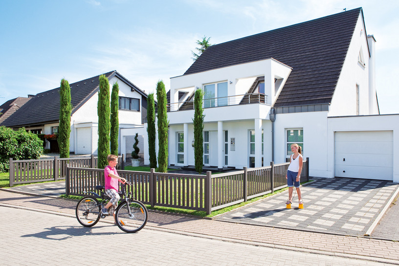 Modernes Einfamilienhaus mit Vorgartenzaun aus WPC, Kind mit Fahrrad und Frau im Einfahrtsbereich.