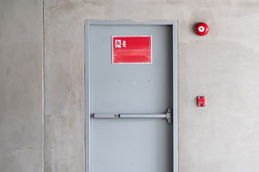 Graue Brandschutztür mit Fluchtwegsymbol und roter Alarmglocke.